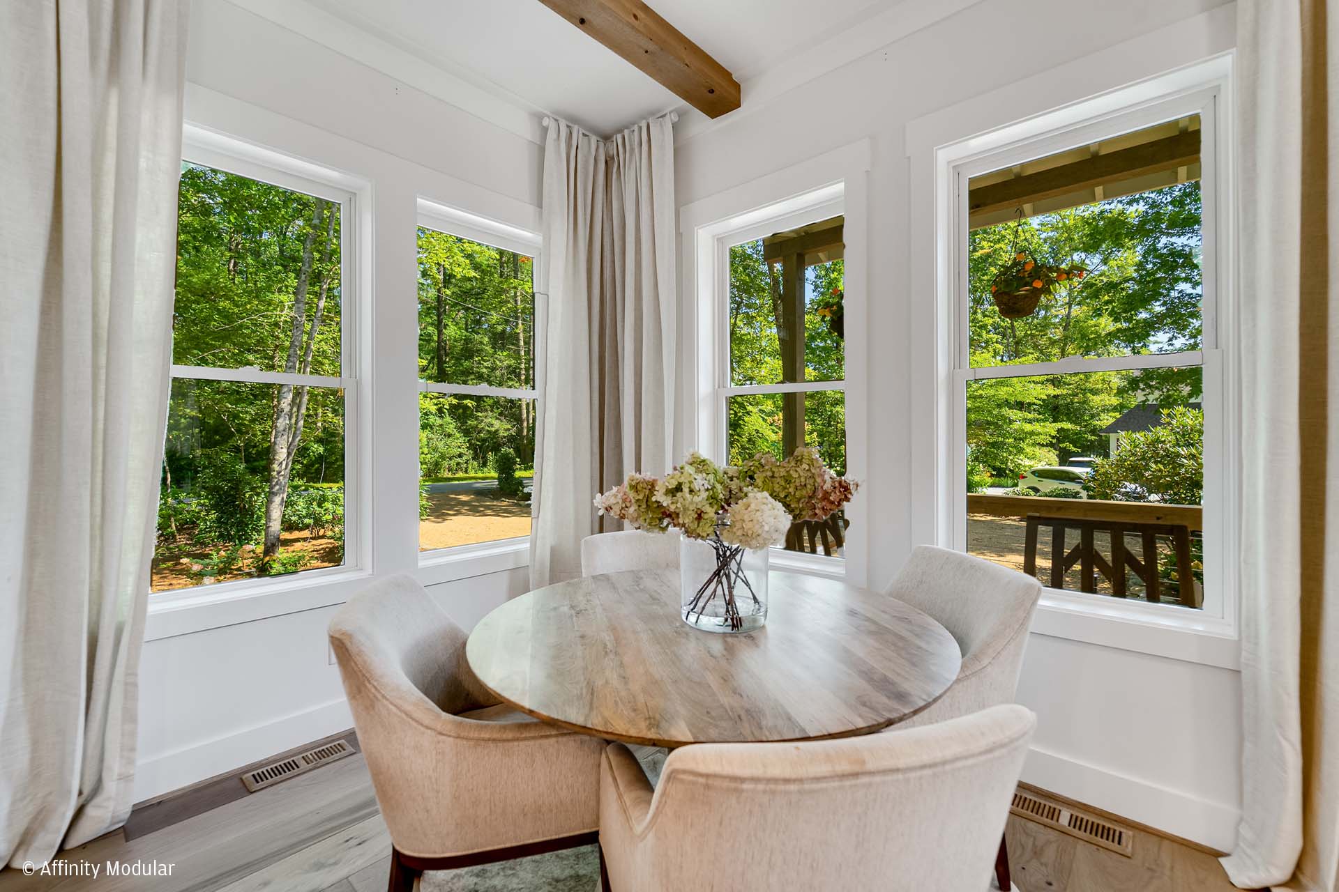 A round table with four chairs by large windows in a modular home, overlooking a green outdoor view with trees.