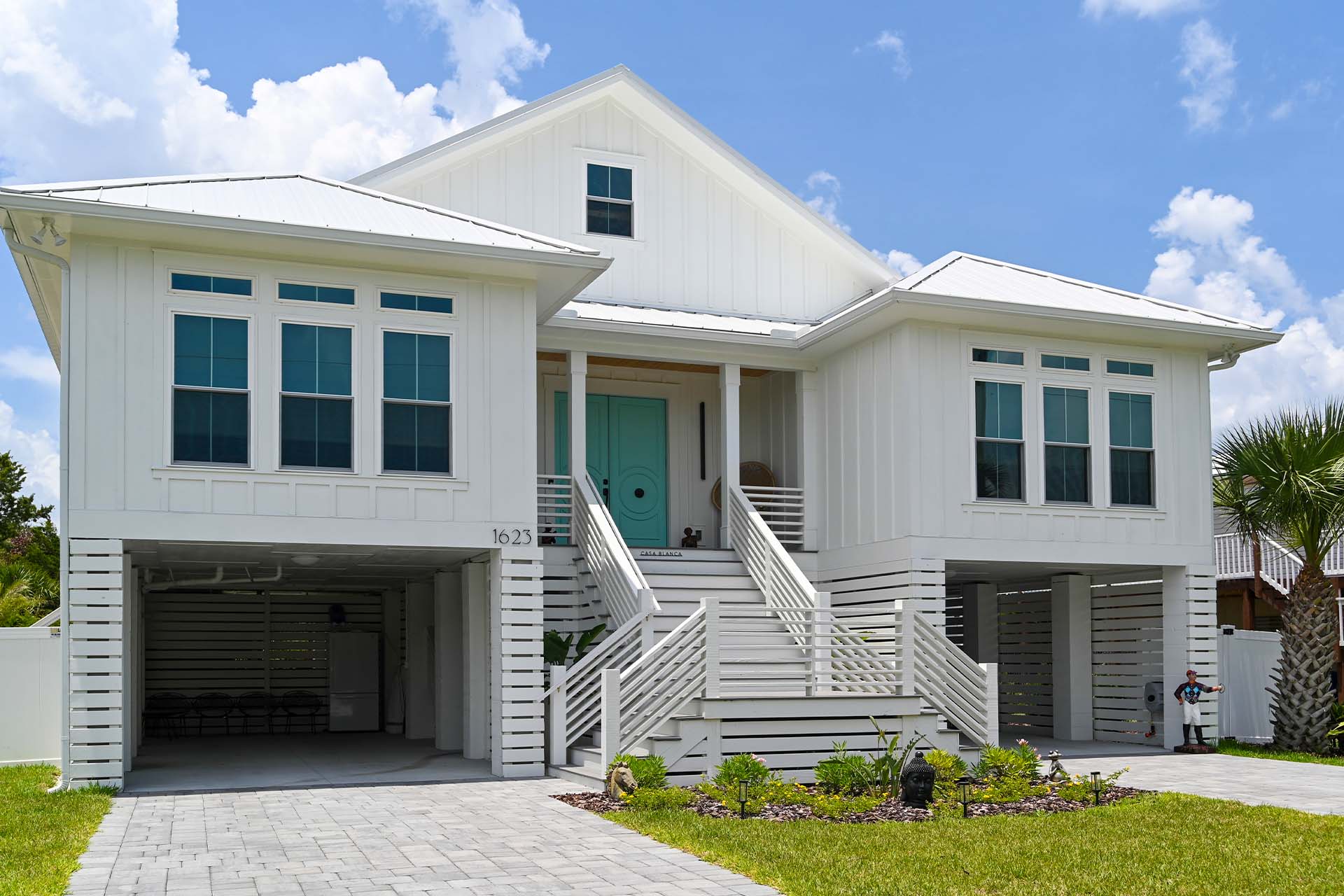 Two-story white modular home with turquoise double doors, large windows, and a spacious driveway under a blue sky.