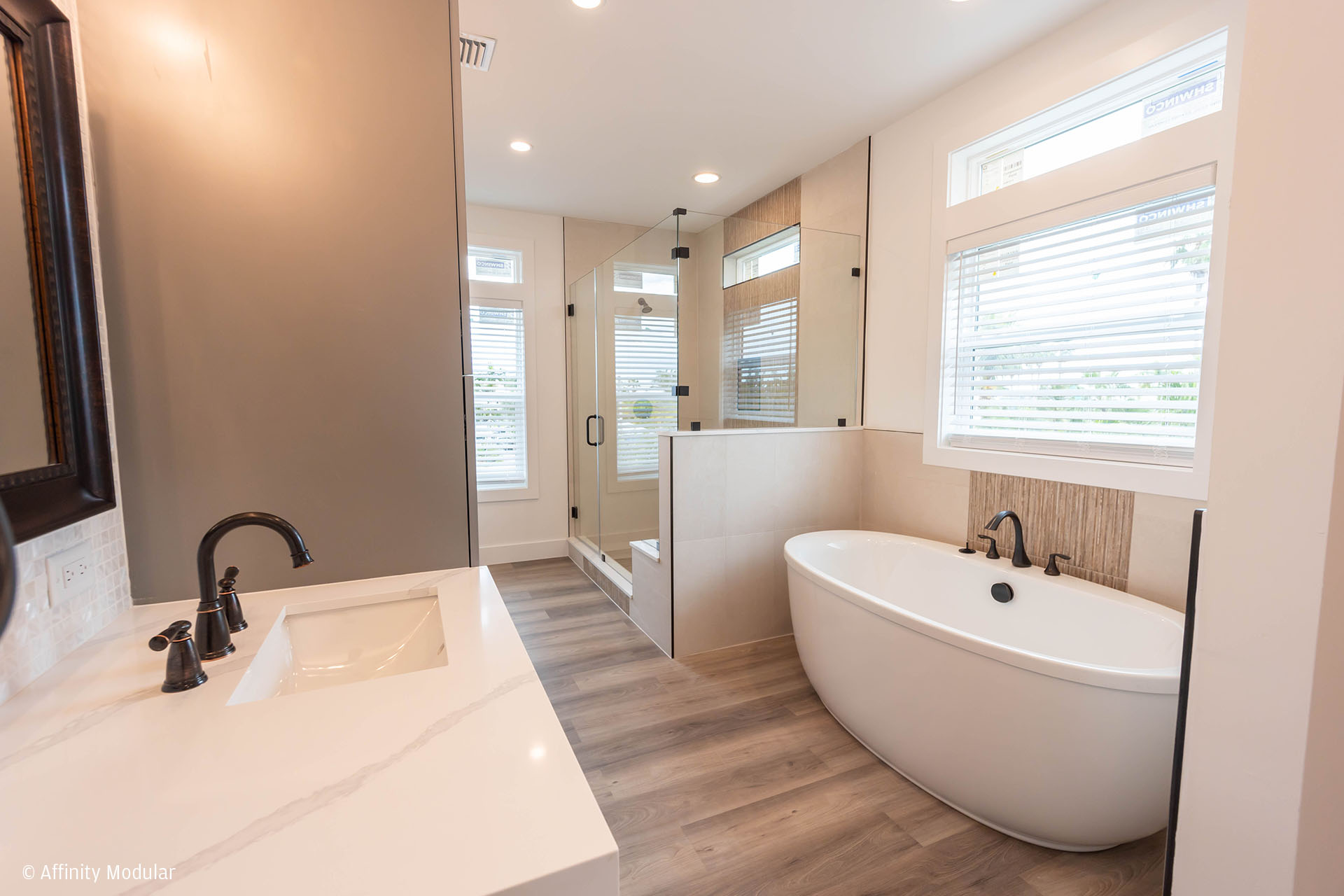 Modern bathroom in a modular home featuring a freestanding tub, glass shower, double sinks, and a large window with blinds.