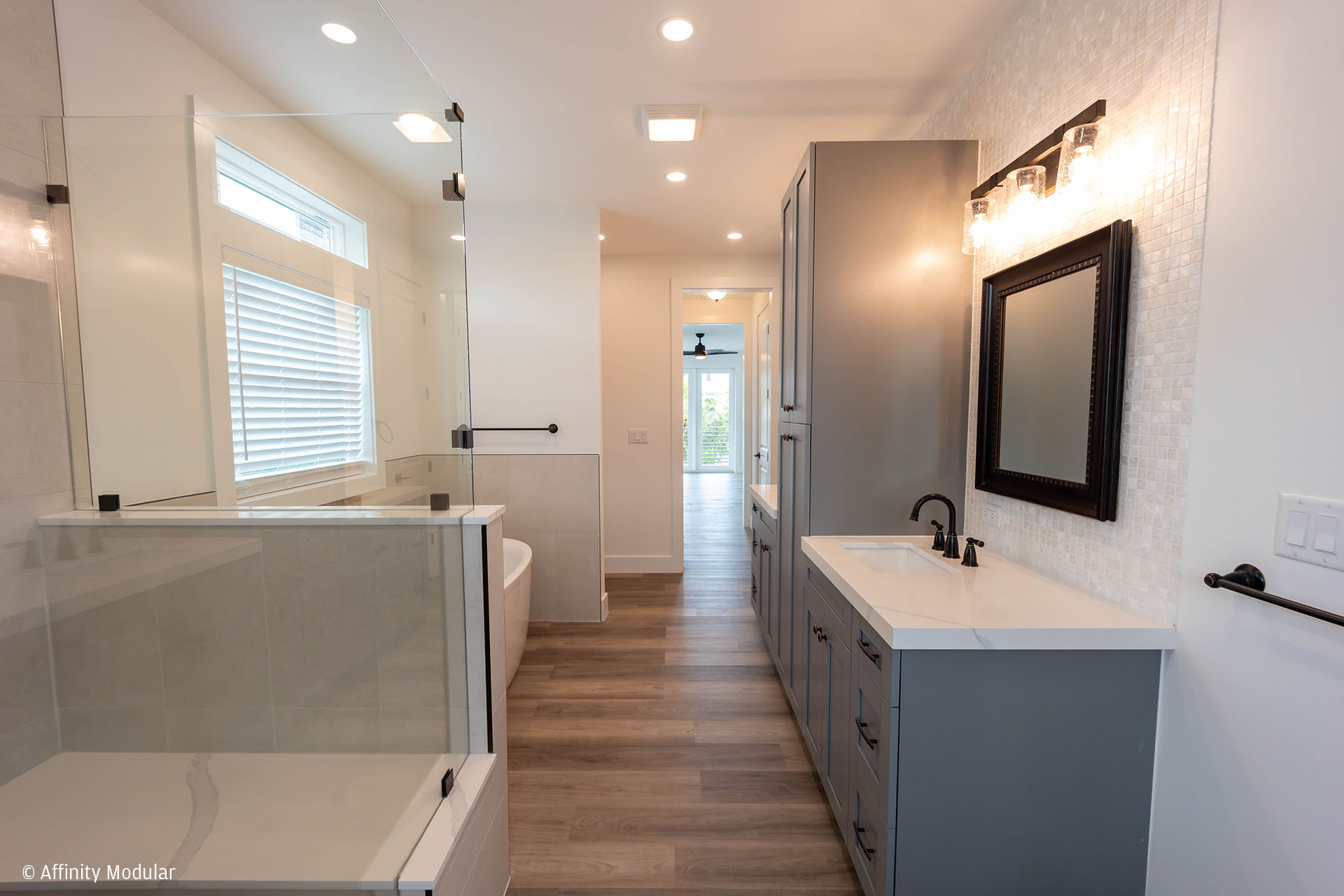 Modern bathroom in a modular home with glass shower, soaking tub, large mirror, gray cabinets, and wood-look flooring.