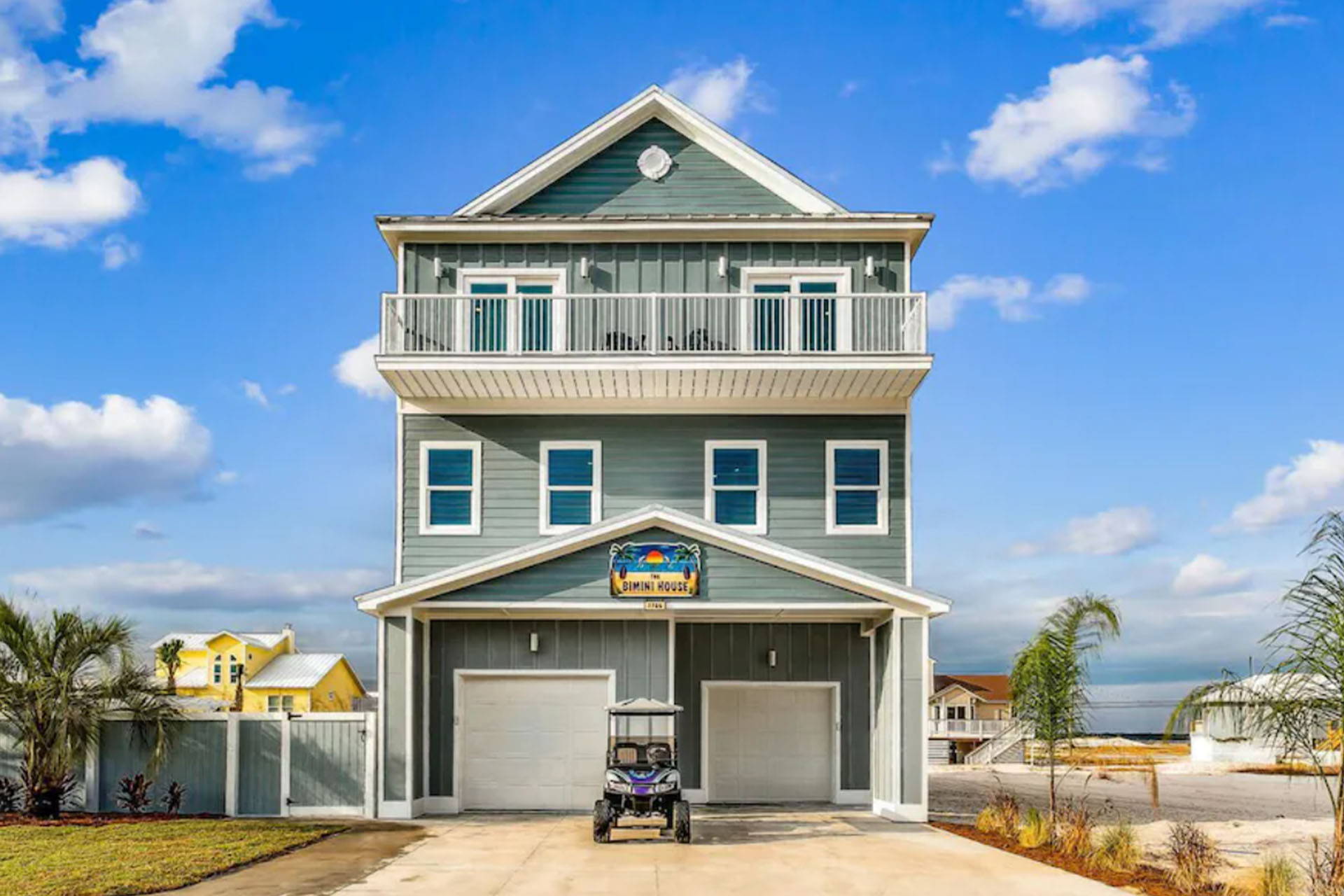 Three-story blue beach house with balconies, a driveway, and a golf cart parked in front, under a blue sky.